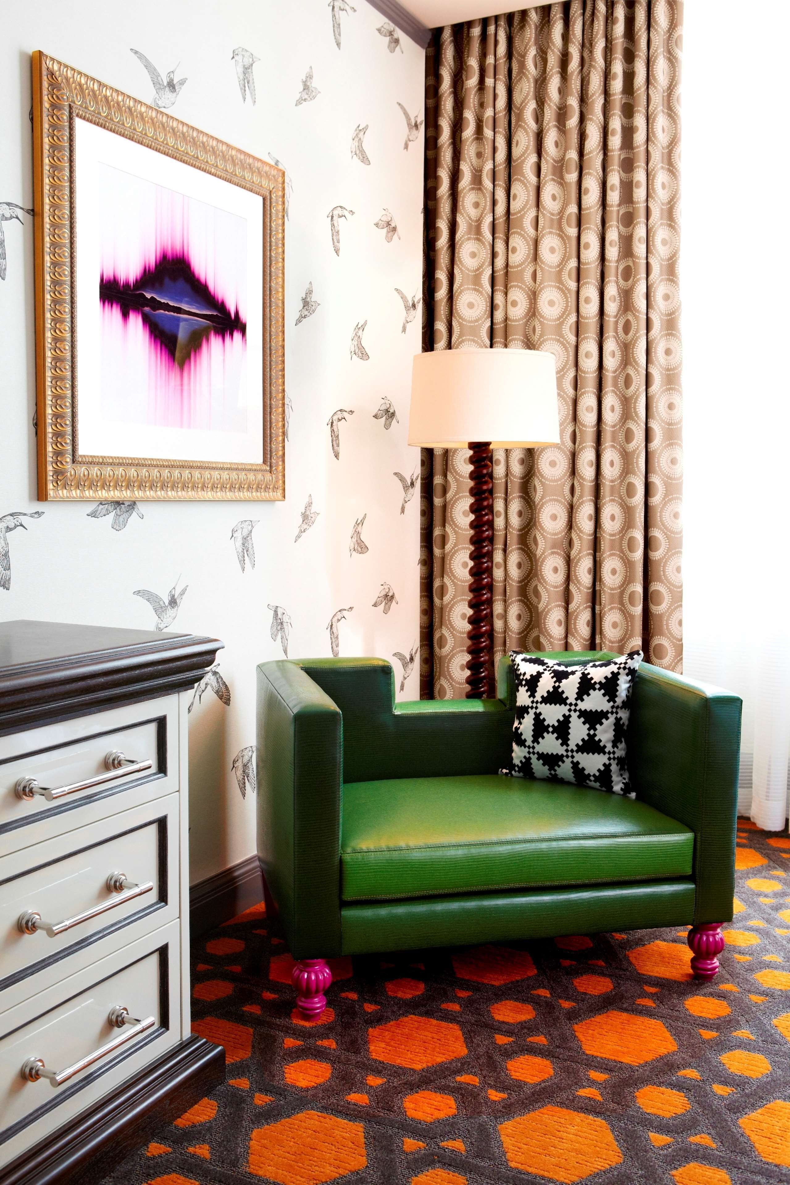 A room with a green armchair, a patterned curtain, a framed artwork, and a patterned carpet.