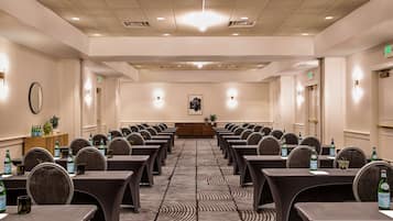Sala de conferências com mesas e cadeiras dispostas em fileiras, equipada com garrafas de água e um pódio na frente.