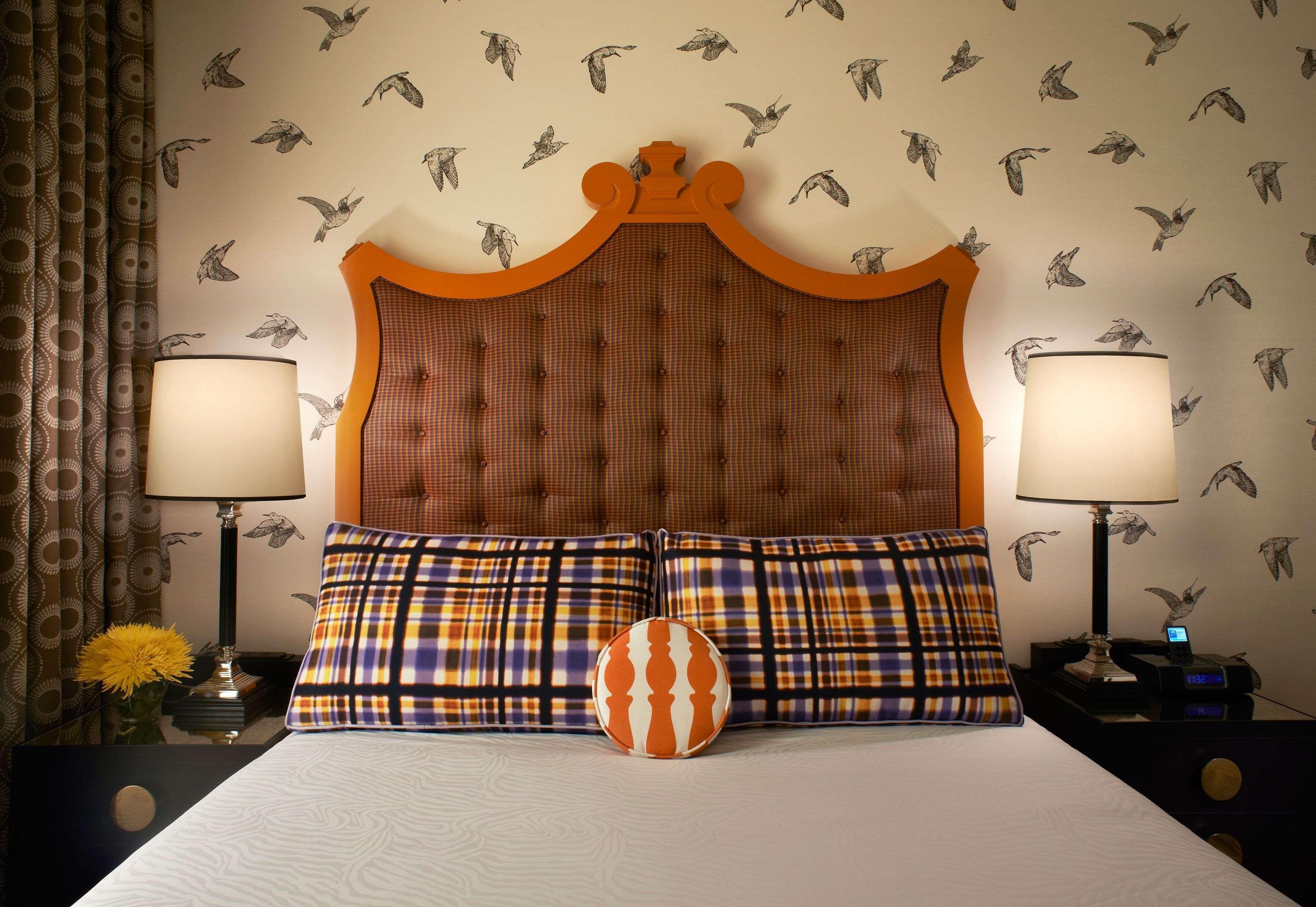 Habitación de hotel con cabecera capitonada, dos lámparas, cama con ropa de cama a cuadros y una pared con diseños de pájaros.