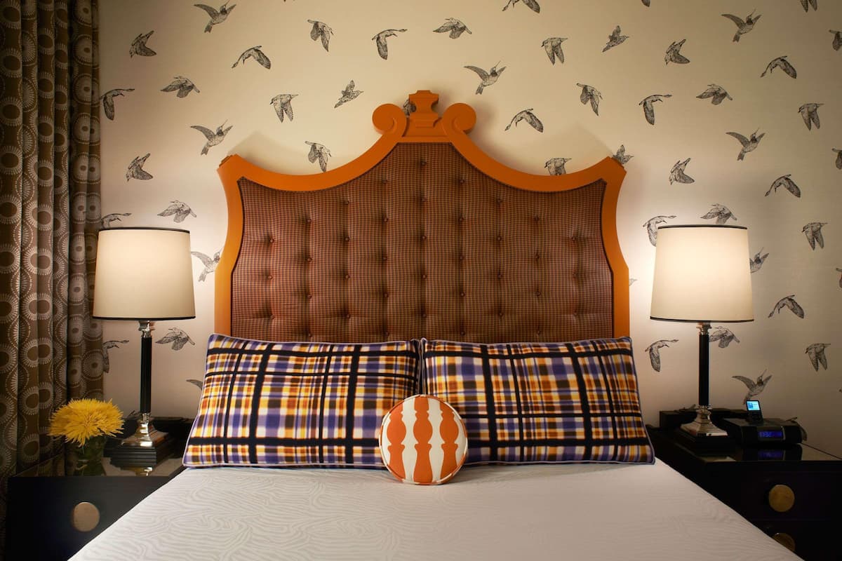 Habitación de hotel con cabecera capitonada, dos lámparas, cama con ropa de cama a cuadros y una pared con diseños de pájaros.