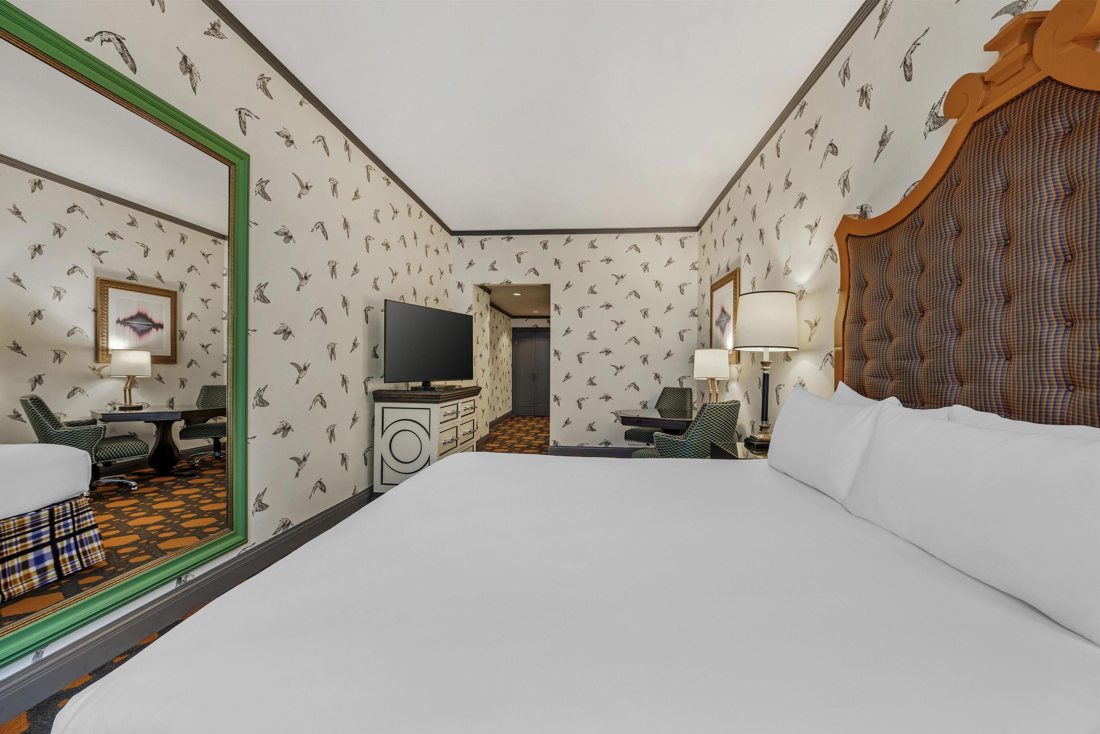 Quarto de hotel com uma cama grande, uma mesa de trabalho com cadeira, televisão e espelho.