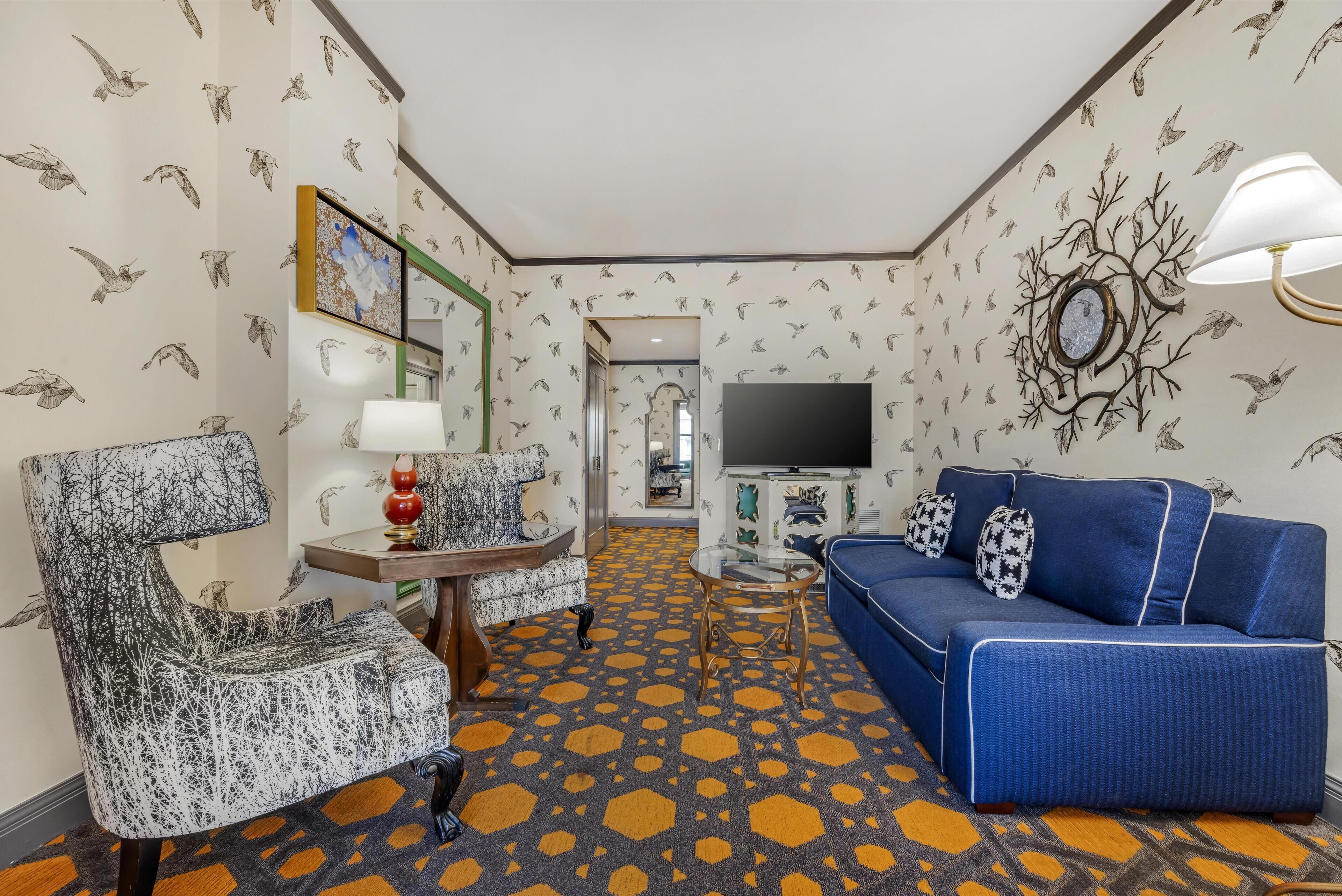 Una habitación de hotel con alfombra estampada, un sofá azul, un sillón estampado, una mesita redonda y un televisor.