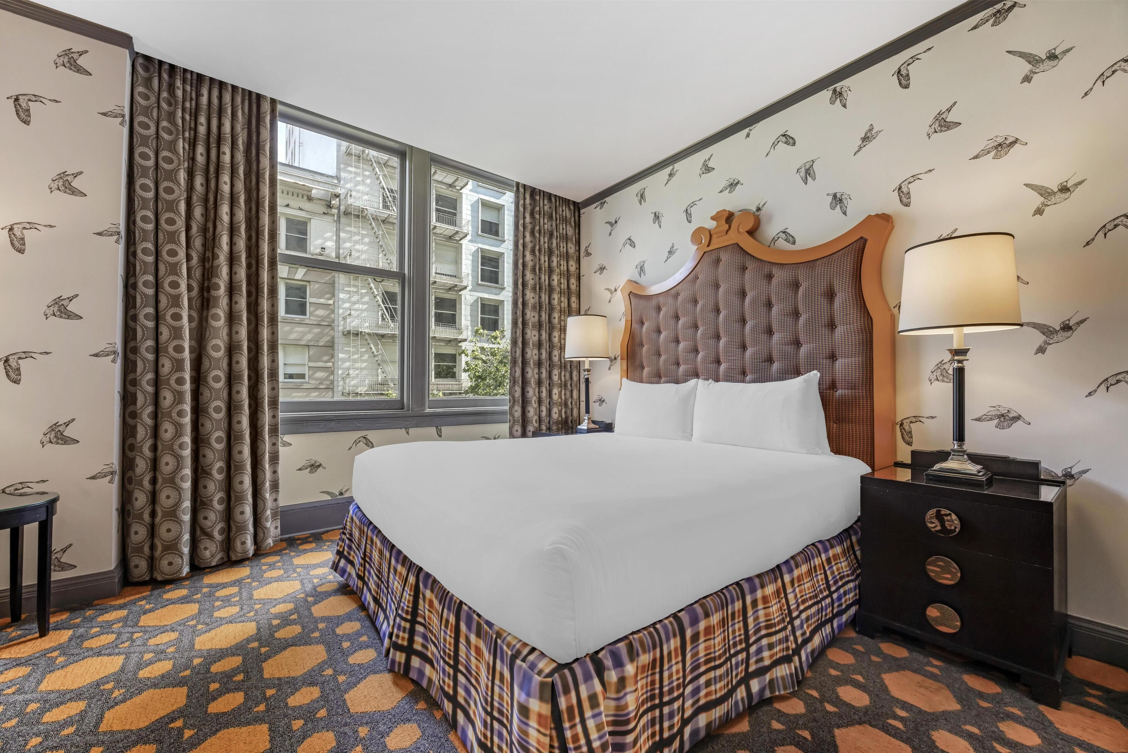 A hotel room with a large bed, a nightstand with a lamp, patterned curtains, and a window with a view of buildings.