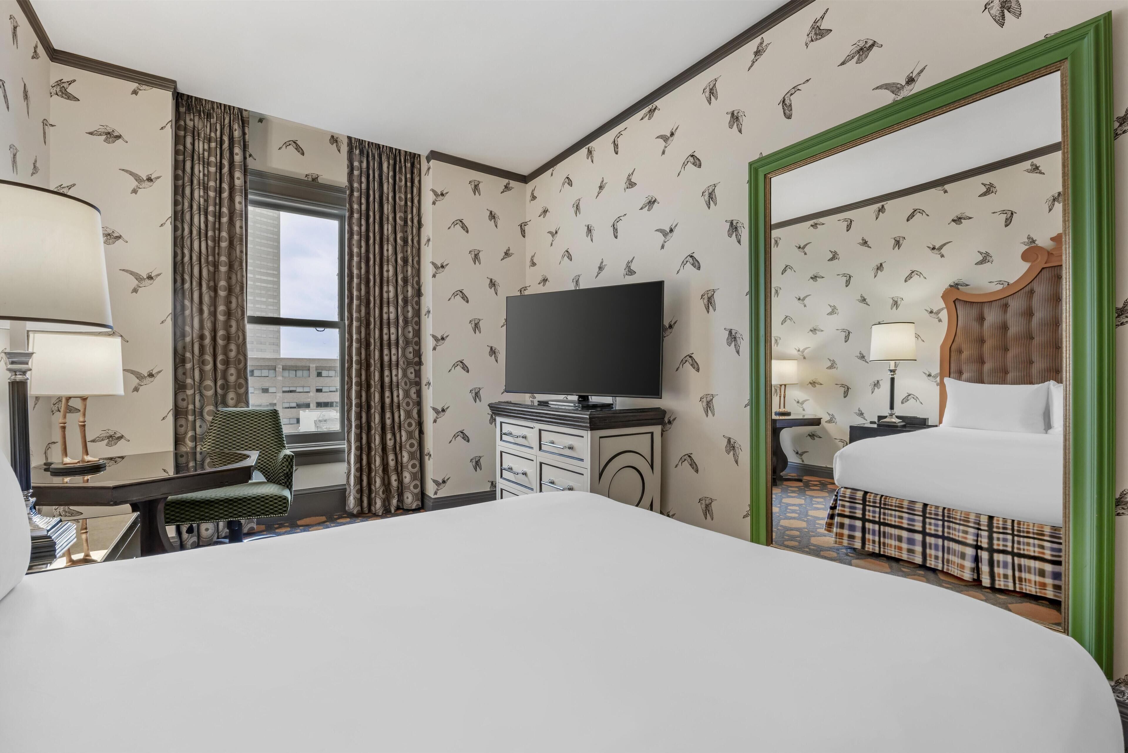 A hotel room with a large bed, a television, a desk with a lamp, a chair, and a nightstand with a lamp.