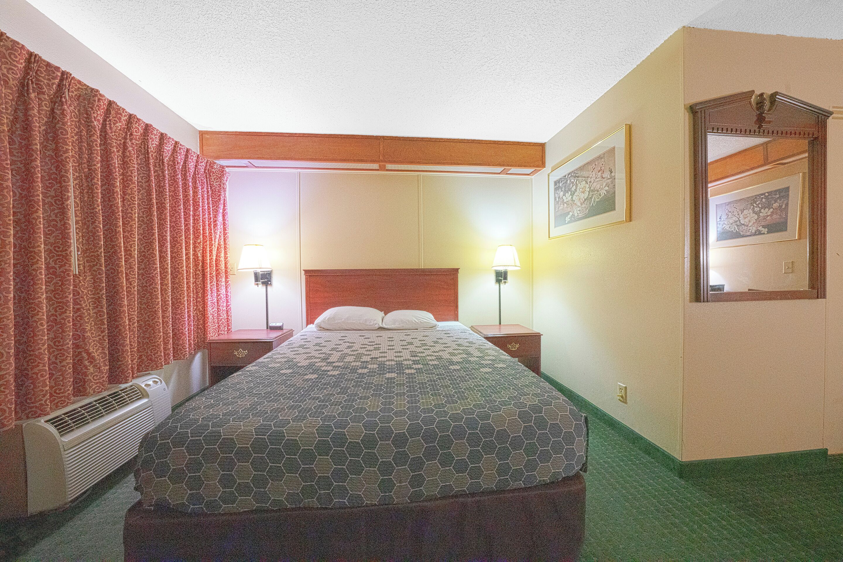 Una habitación de hotel con cama, dos lámparas de noche, un calefactor, cabecera de madera, espejo y alfombra verde.
