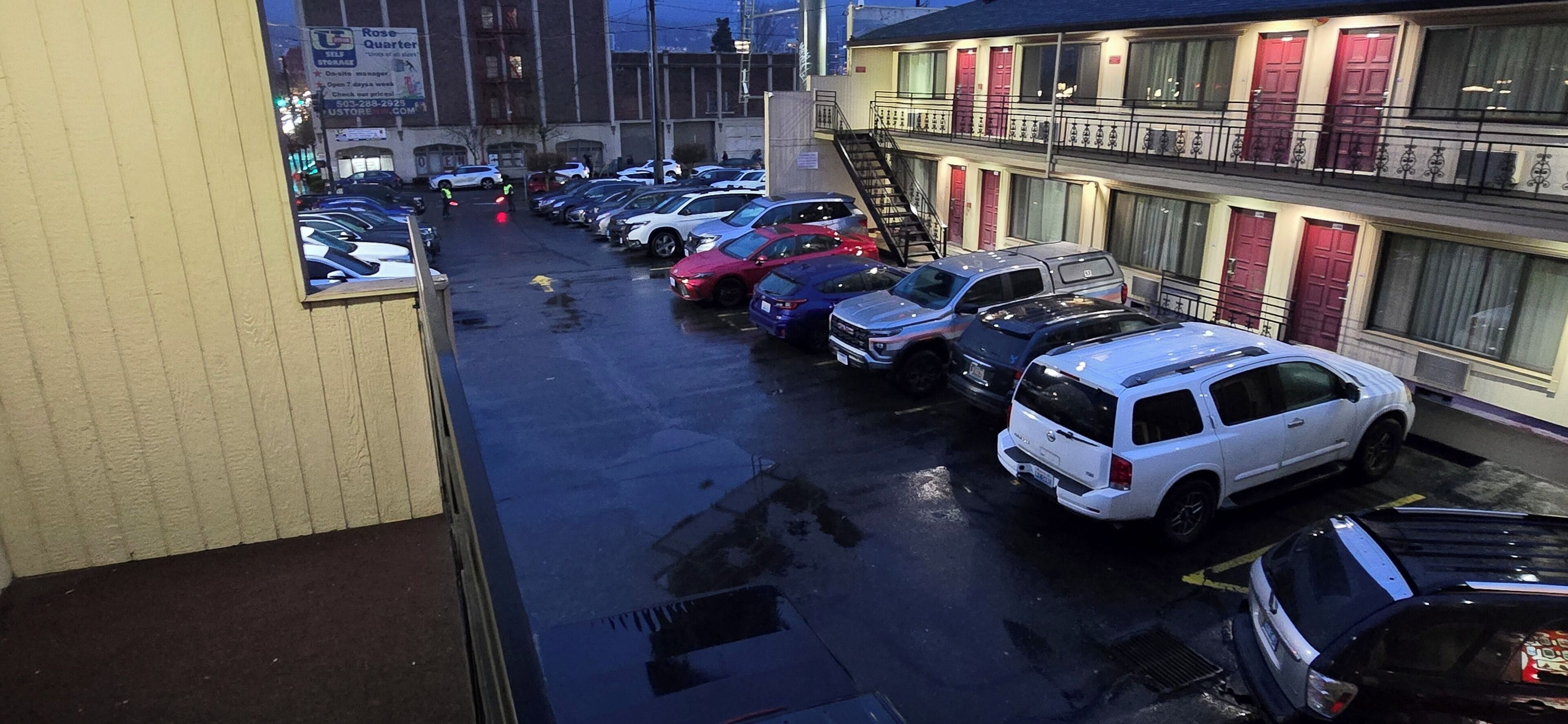 Ein Parkplatz mit mehreren in Reihen parkenden Autos, neben einem Gebäude mit roten Türen und einer Treppe, die zu einer oberen Etage führt.
