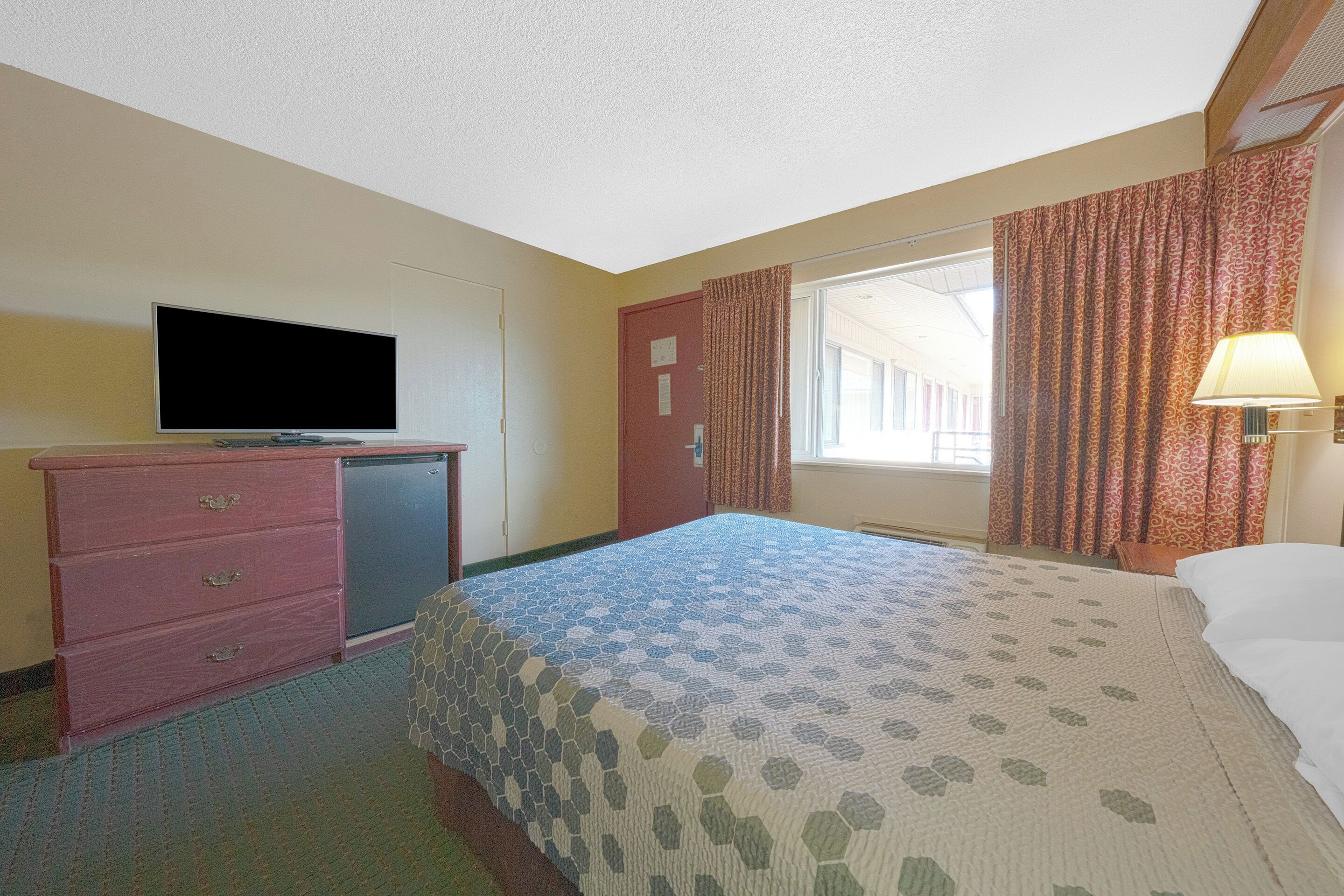 A hotel room with a bed, a television on a dresser, a window with curtains, and a lamp.