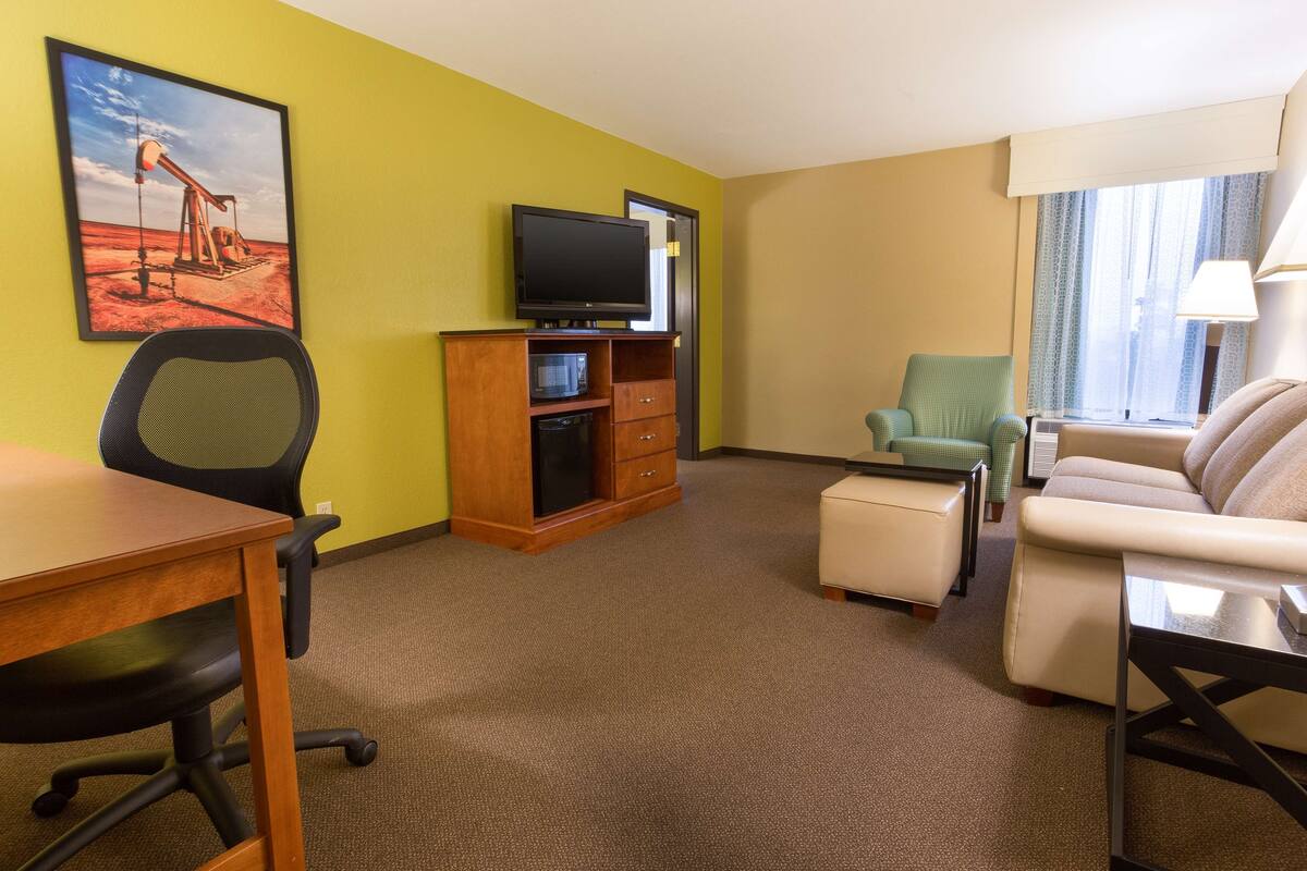 A hotel room with a wooden desk, a television on a wooden stand, a green armchair, a beige sofa, and a window with curtains.