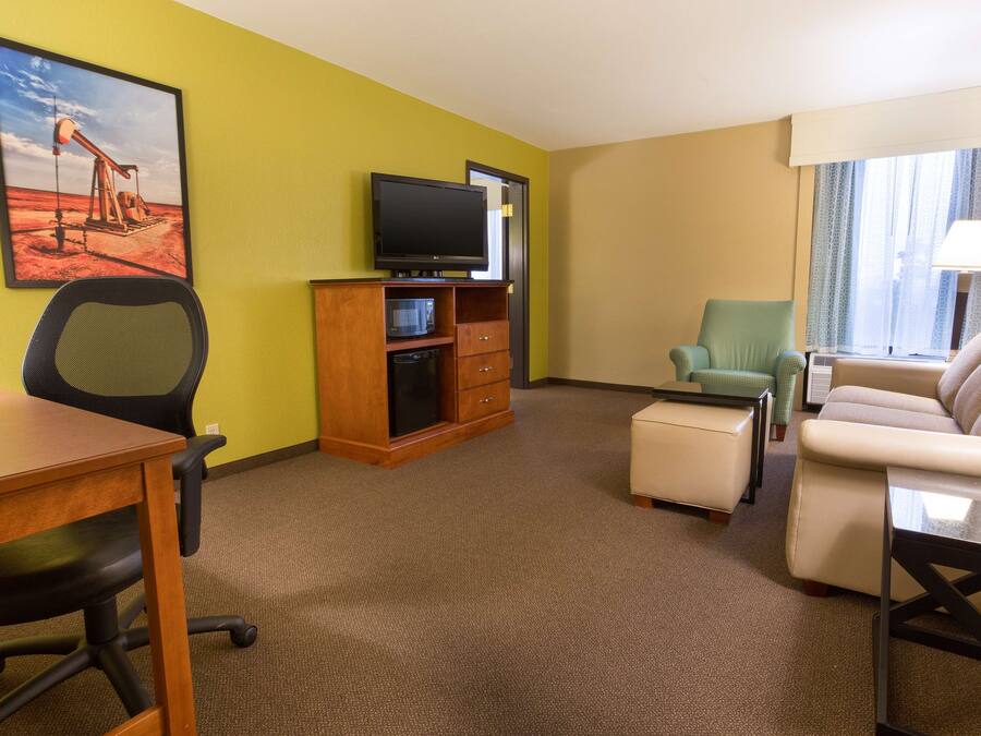 A hotel room with a wooden desk, a television on a wooden stand, a green armchair, a beige sofa, and a window with curtains.