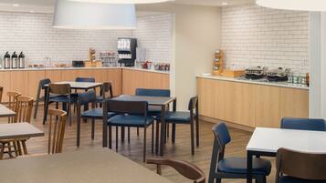 A modern cafe with wooden tables and chairs, a counter with coffee machines, and a white brick wall.