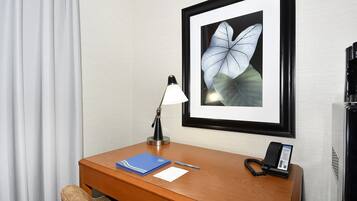 A hotel room with a wooden desk, a framed butterfly artwork, a desk lamp, a telephone, a notepad, and a pen.