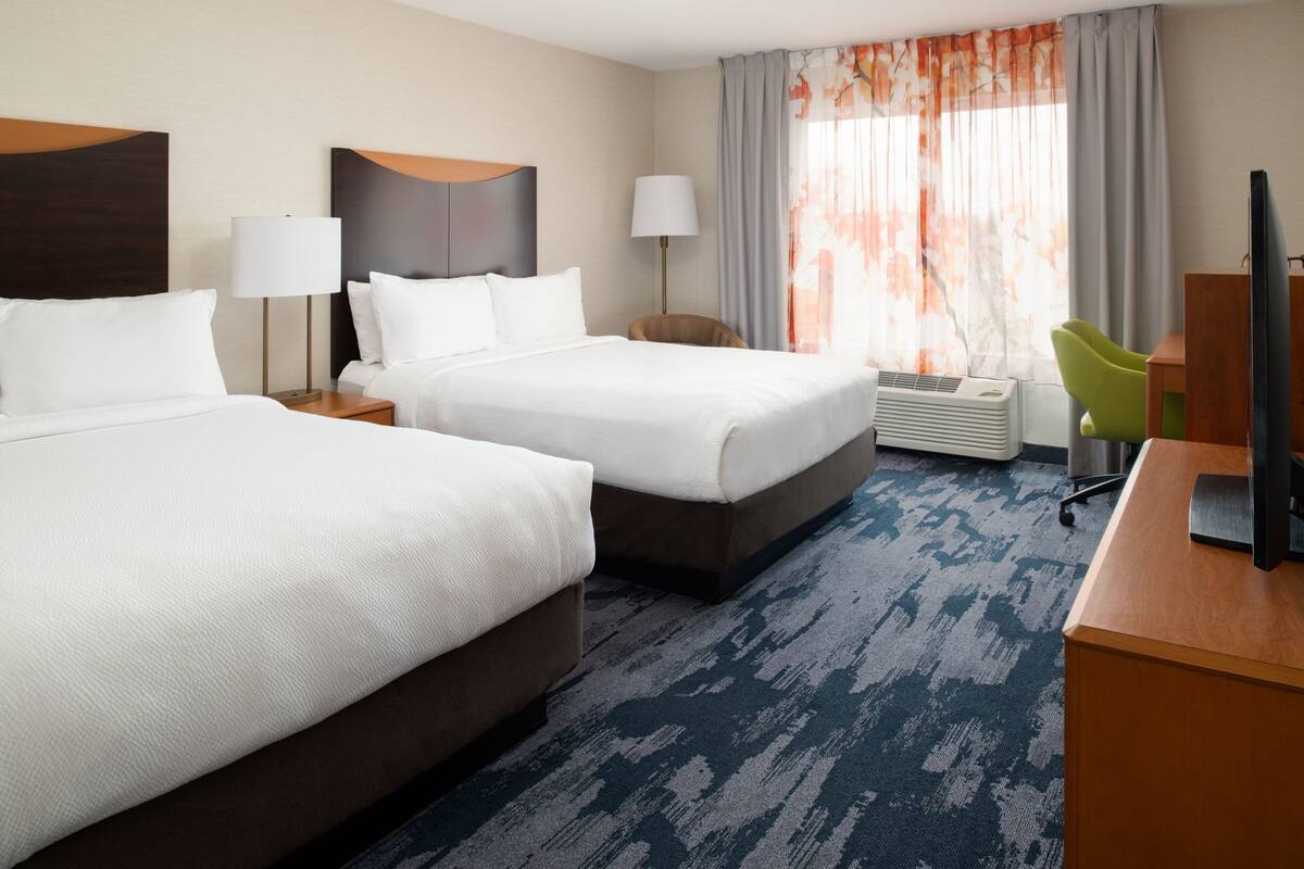 A hotel room with two beds, a desk, a chair, a TV, and a window with curtains.