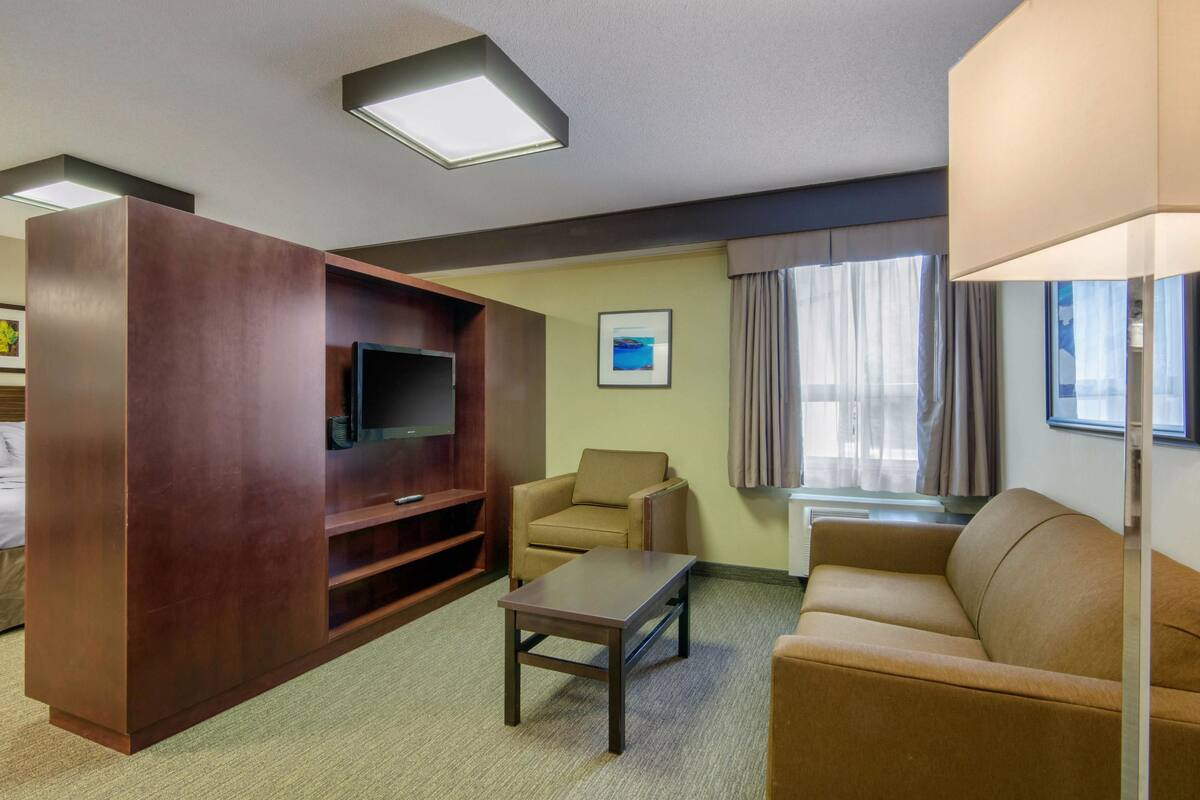 Une chambre d’hôtel avec une grande armoire en bois, un téléviseur à écran plat, un canapé beige, une petite table et une fenêtre avec des rideaux.