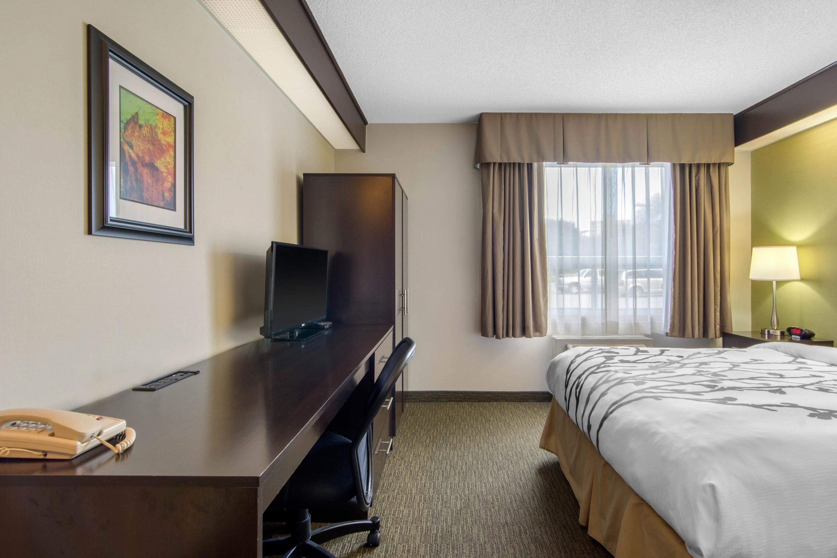 Quarto de hotel com cama, escrivaninha com televis ão, cadeira, abajur e janela com cortinas.