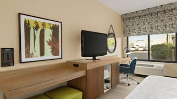 A hotel room with a bed, a desk with a television, a green chair, a large window with a view, and a framed picture on the wall.