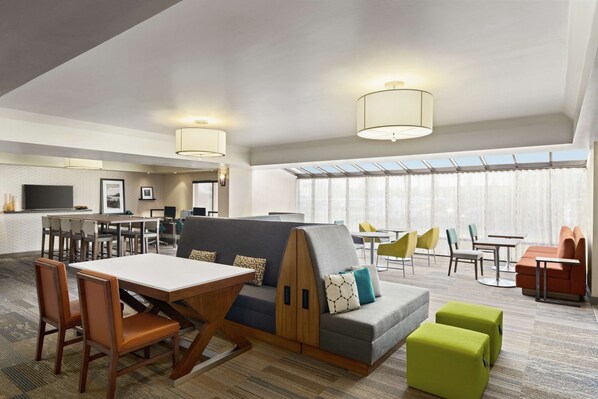 A modern hotel lobby with a dining area, seating arrangements, and a large window providing natural light.