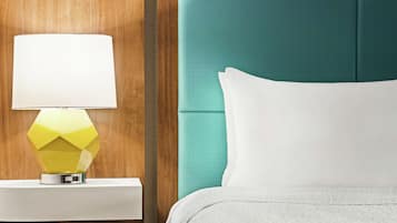 A hotel room with a teal headboard, a white bedspread, and a geometric yellow lamp on a white nightstand.