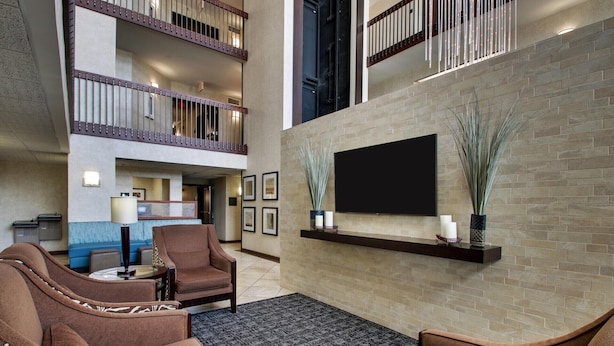 A spacious lobby with a high ceiling, a flat-screen TV mounted on a brick wall, a seating area with brown upholstered chairs, and a staircase leading to an upper level. - Sonesta Essential Houston Hobby Airport (Houston)