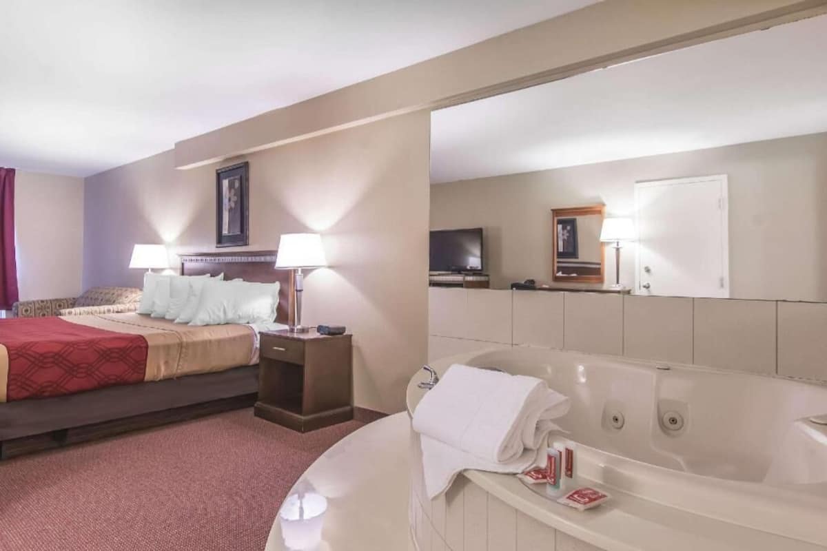 A hotel room with a bed, bedside lamps, a TV, a bathroom with a bathtub, and a mirror.