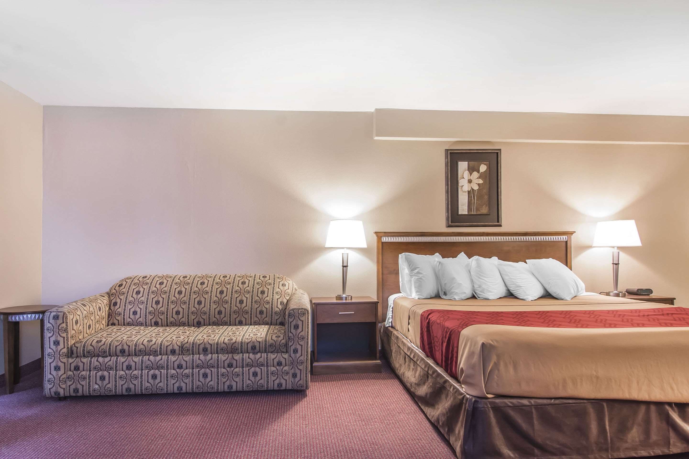 Ein Hotelzimmer mit einer gemusterten Couch, einem Bett mit roter Decke, zwei Nachttischen mit Lampen und einem gerahmten Bild an der Wand.