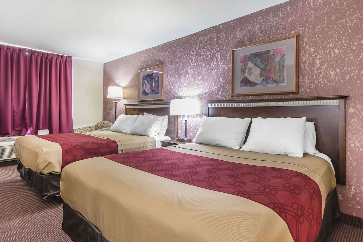 A hotel room with two beds, each with a headboard, a red patterned bedspread, and white pillows. There are two bedside lamps, a framed picture on the wall, and a window with curtains.