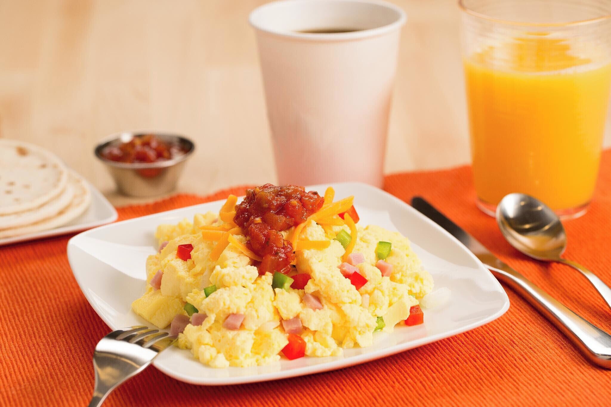 A breakfast plate with scrambled eggs, ham, and vegetables, a cup of coffee, and a glass of orange juice on a table.