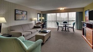 A hotel room with a sofa, armchair, dining table, chairs, a TV, and a large window with curtains. - Heritage Inn & Suites The Woodlands, Trademark by Wyndham (The Woodlands)