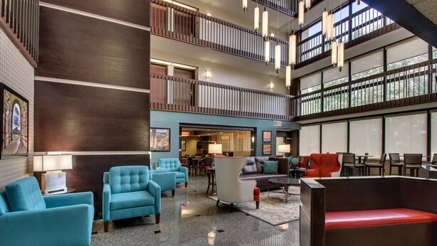 A spacious lobby with multiple seating areas, including blue armchairs and a red bench, and a view of the upper floor through large windows. - Heritage Inn & Suites The Woodlands, Trademark by Wyndham (The Woodlands)