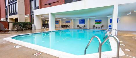 Una piscina interior con agua azul cristalina, rodeada por una solana blanca, y contigua a un edificio con fachada de ladrillo y ventanas amplias.
