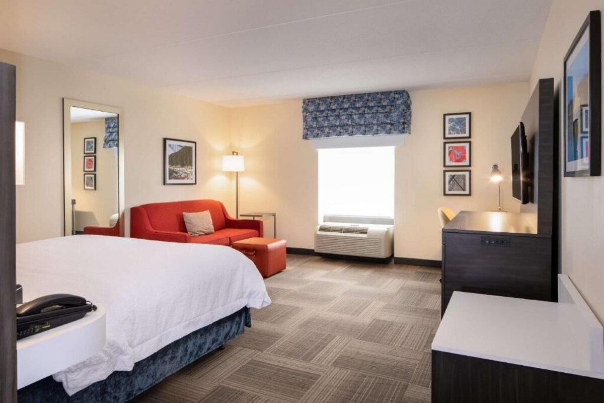 A hotel room with a bed, a red sofa, a desk, a lamp, and a TV.