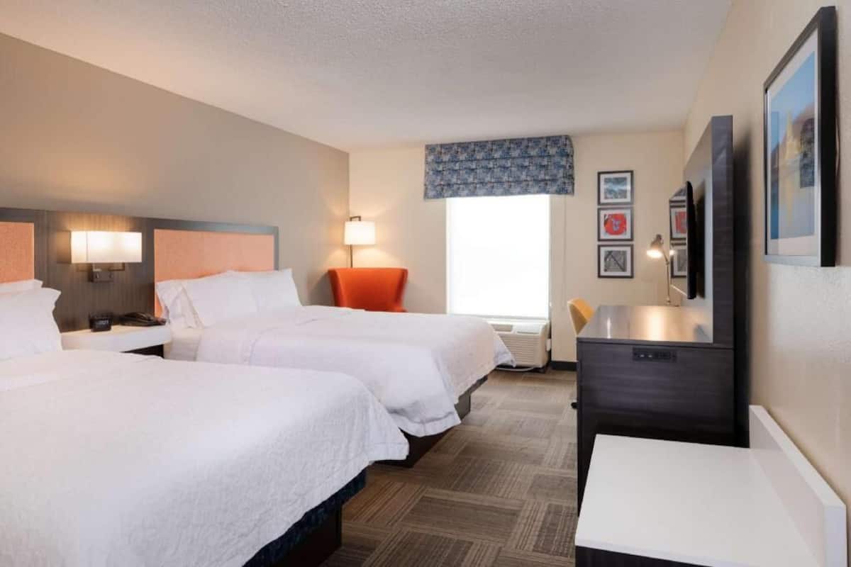 A hotel room with two beds, a desk, a chair, a TV, and a window with a view.