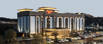 Un hotel Hampton Inn con fachada moderna, amplias ventanas y un estacionamiento lleno de coches.