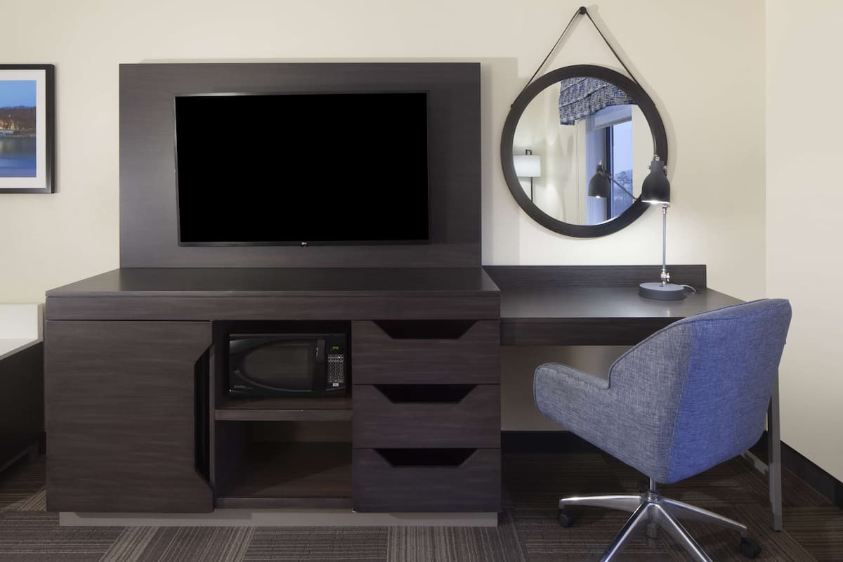 Ein modernes Hotelzimmer mit einem Flachbildfernseher, der an einem dunklen Holztisch befestigt ist, einer Mikrowelle, einem blauen Bürostuhl und einem runden Spiegel an der Wand.
