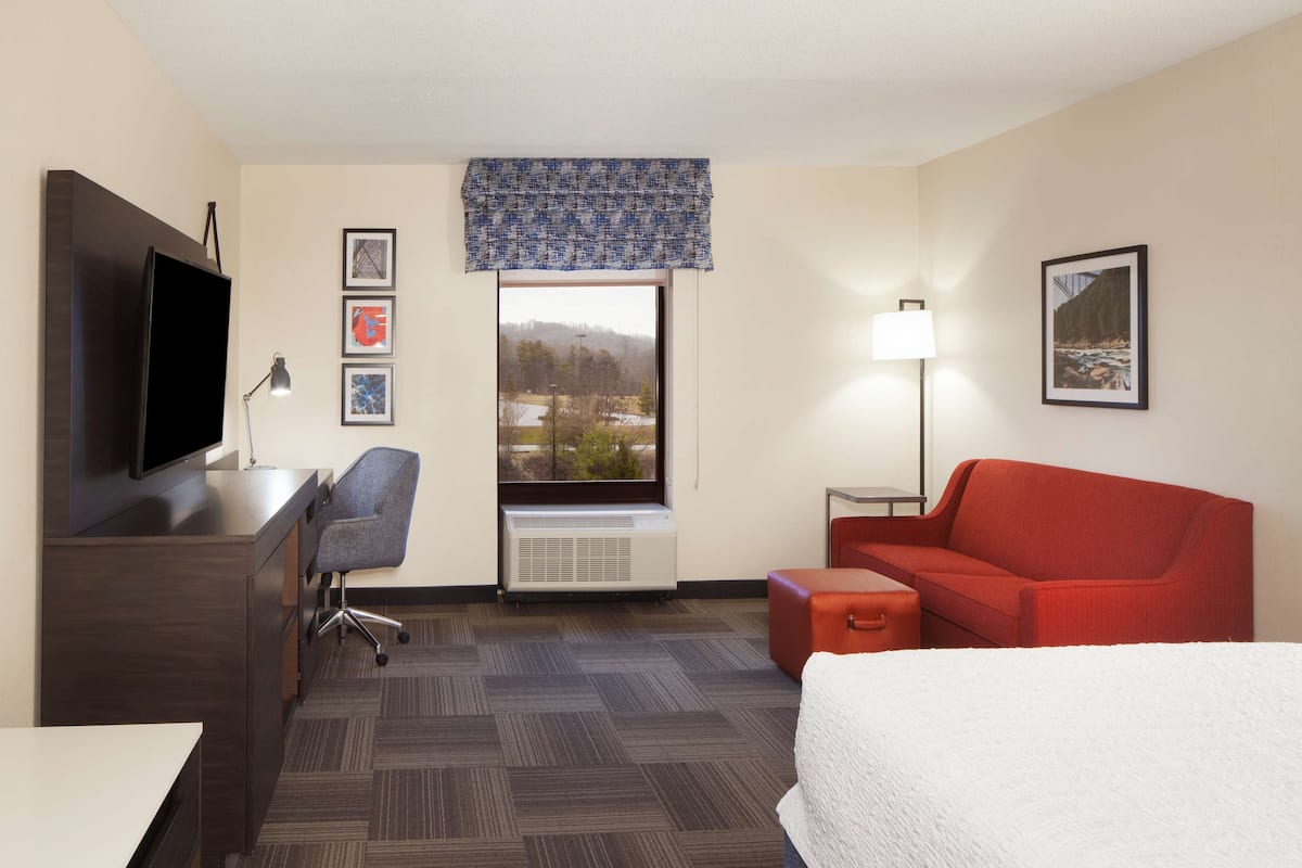 Ein Hotelzimmer mit einem Bett, einem Schreibtisch mit Fernseher, einem Sessel, einem roten Sofa, einem kleinen Tisch, einer Lampe und einem Fenster mit Blick auf eine Landschaft.