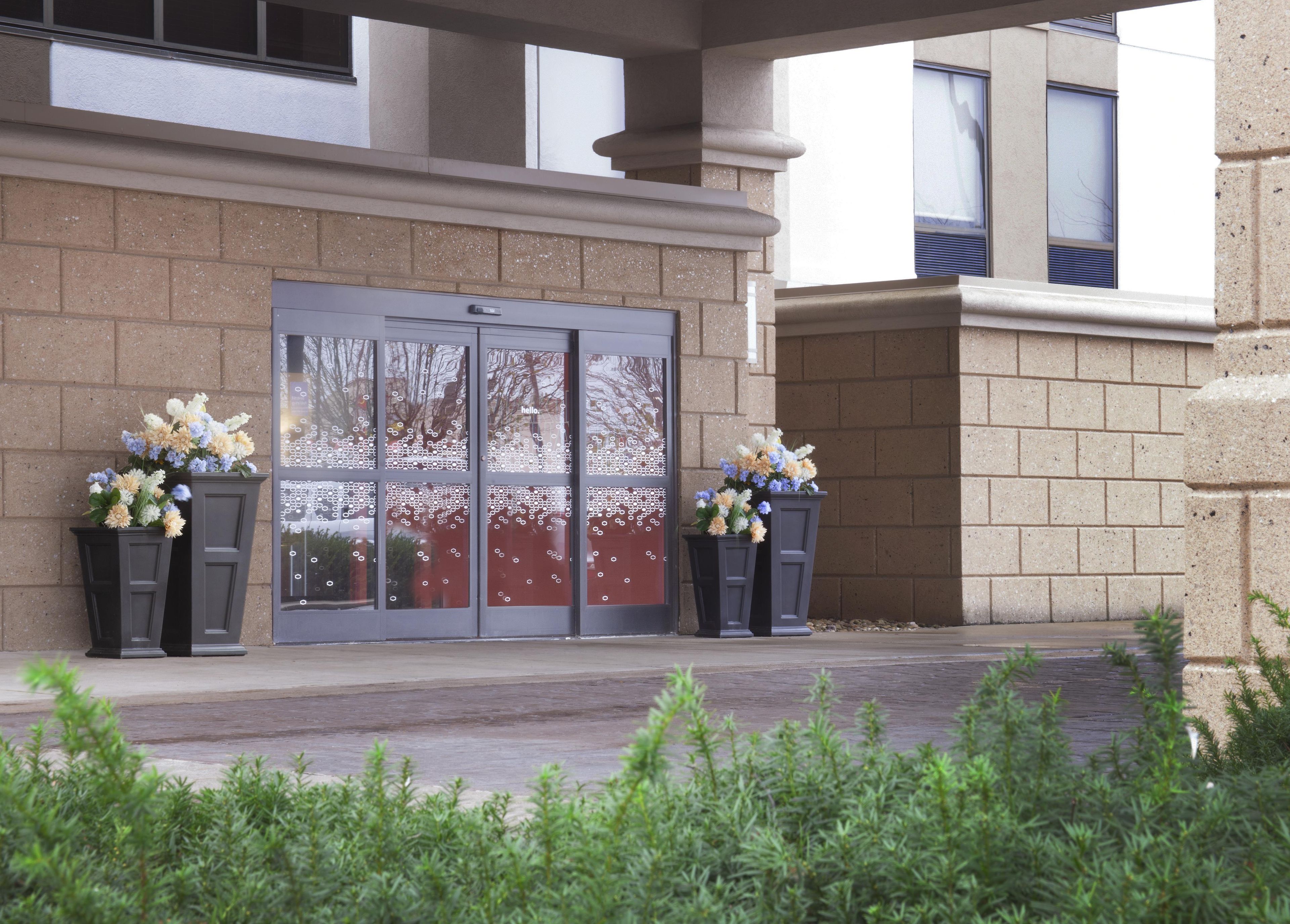 A building entrance with a glass door, two flower arrangements, and a brick wall.