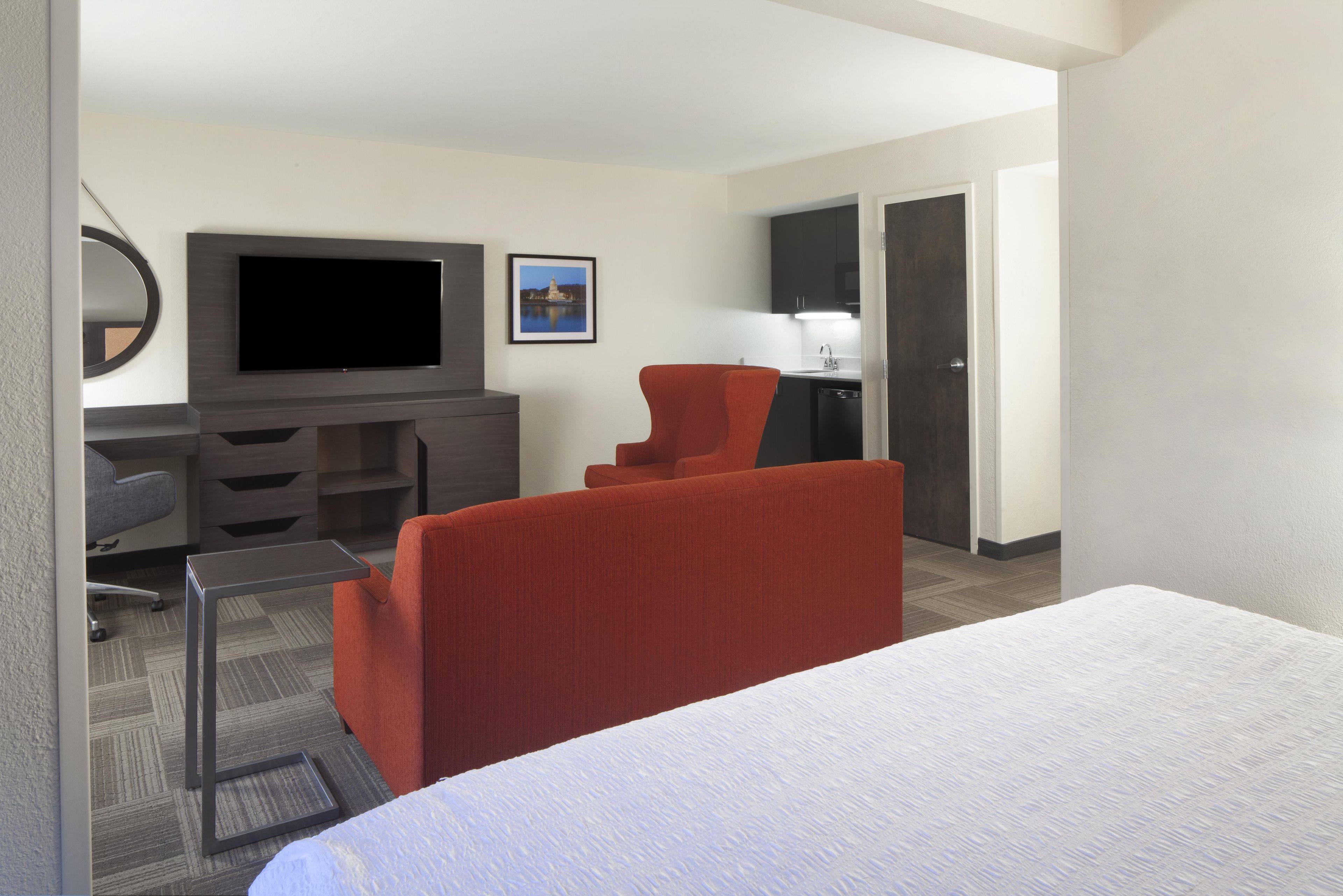 A hotel room with a bed, a desk with a flat-screen TV, a red chair, a small table, and a kitchenette.