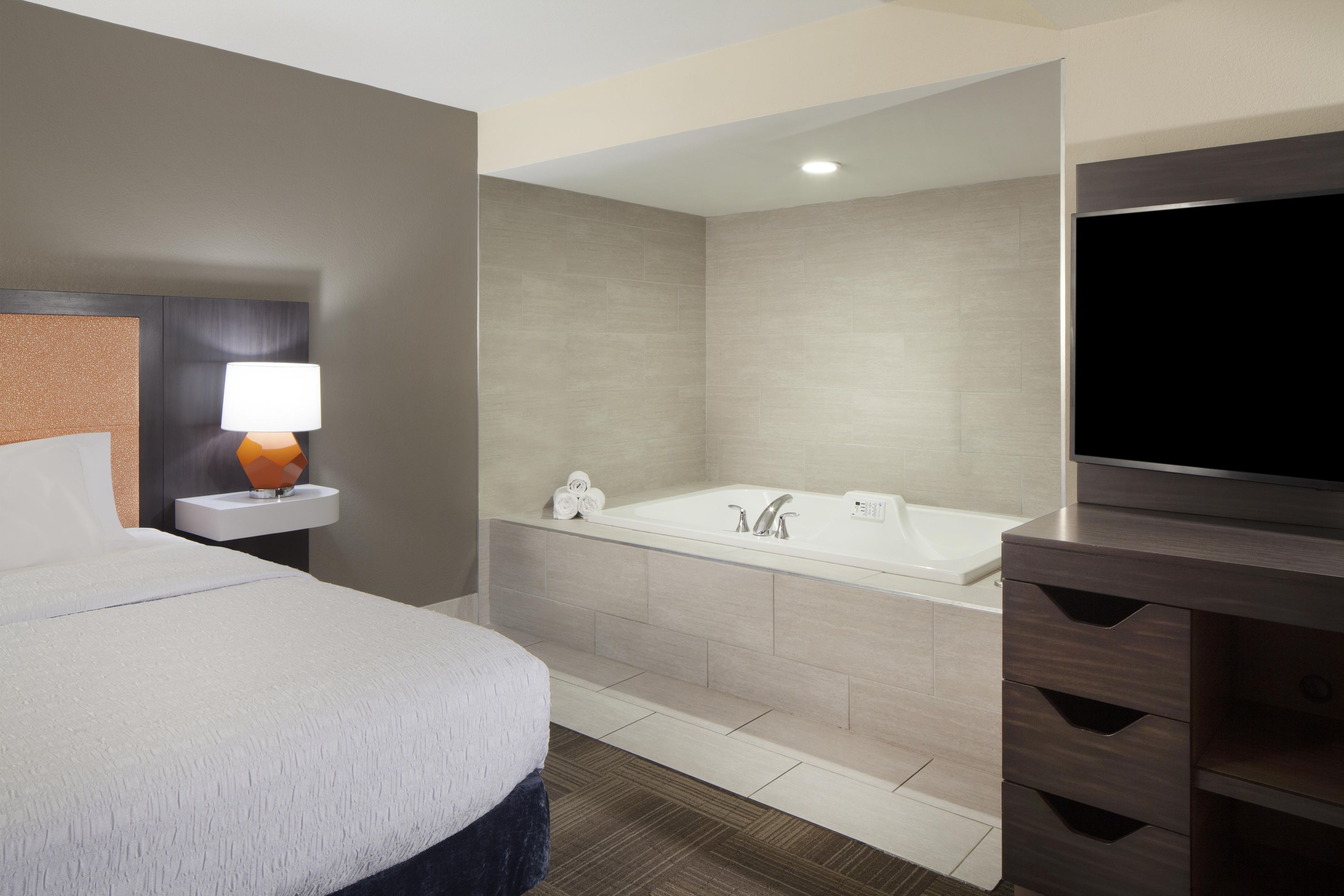 A hotel room with a bed, a bathroom with a bathtub, a TV, and a lamp.