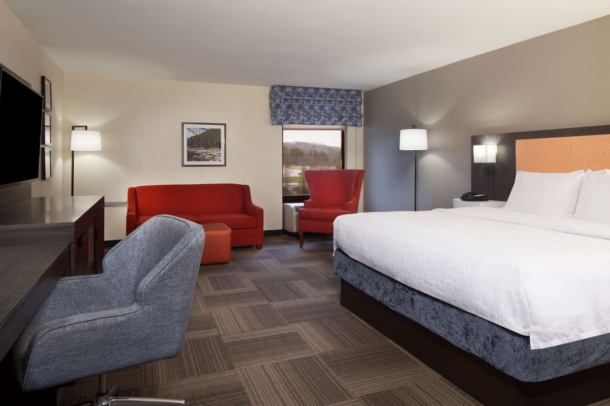 Ein Hotelzimmer mit Bett, Schreibtisch, einem roten Sofa, einem grauen Sessel und einem Fenster mit Blick.