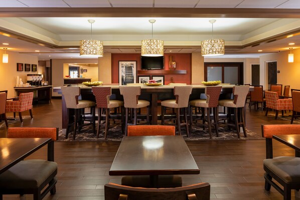 A modern dining area with a central table surrounded by chairs, overhead lighting, and a kitchen in the background. - Hampton Inn Charleston-Southridge (Charleston)