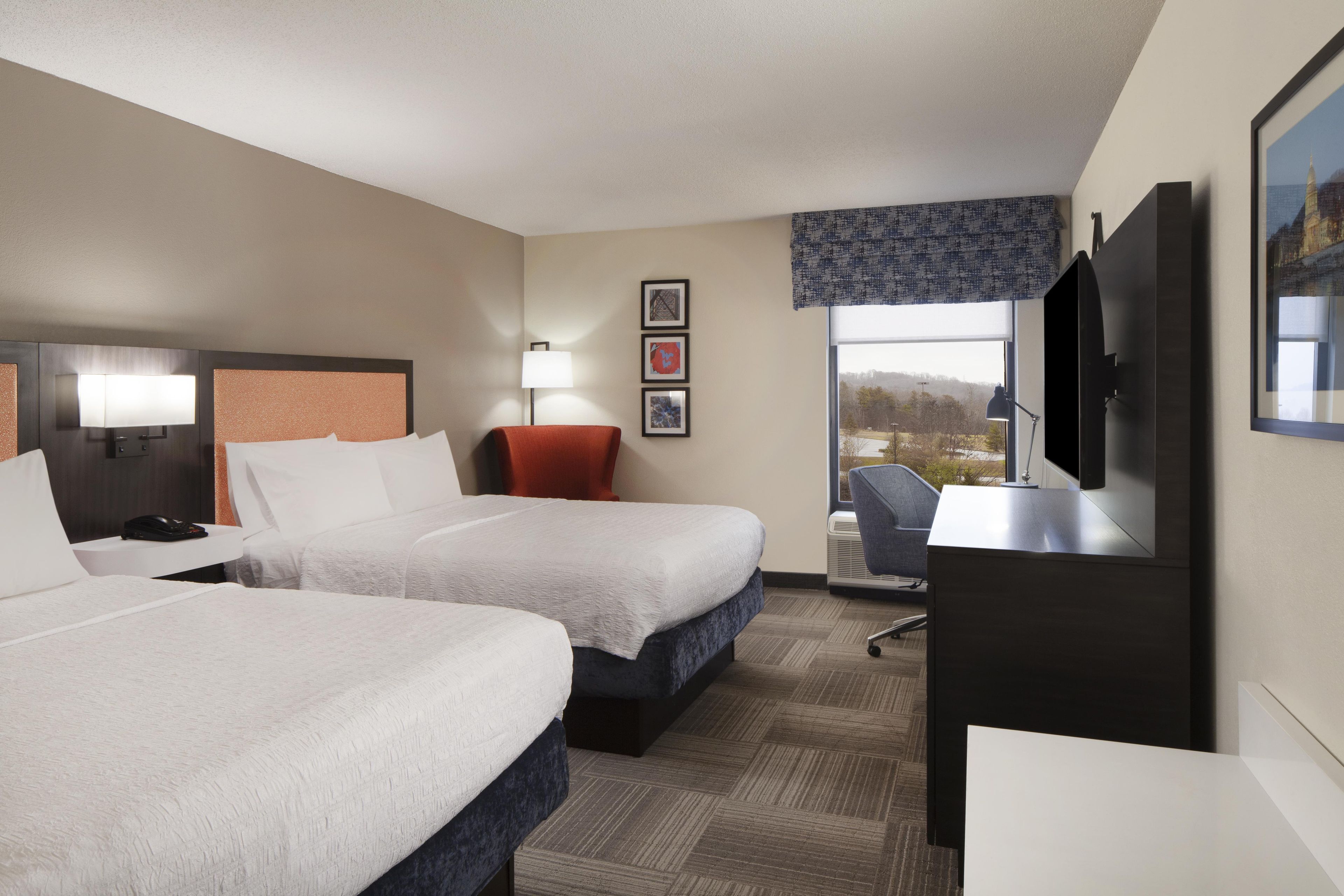 Quarto de hotel com duas camas, uma escrivaninha, uma cadeira, televisão e janela com vista.