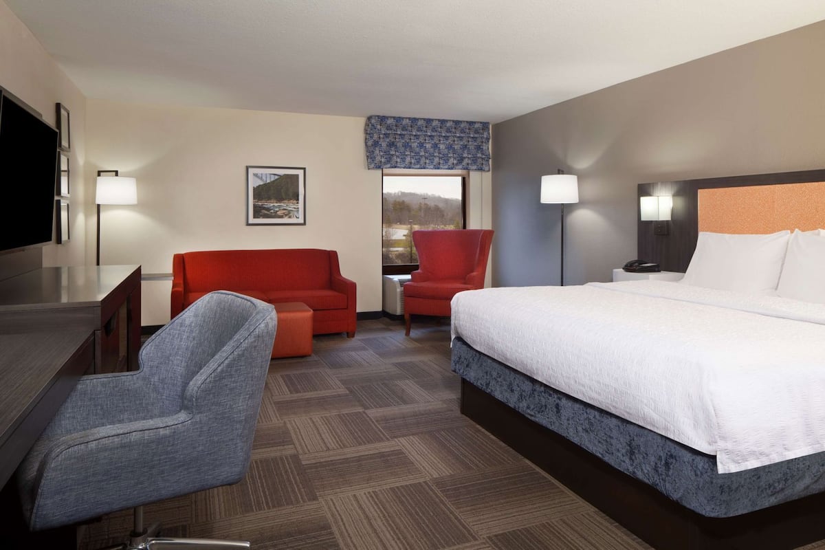A hotel room with a bed, a desk, a TV, a red armchair, a grey armchair, and a window with a view.