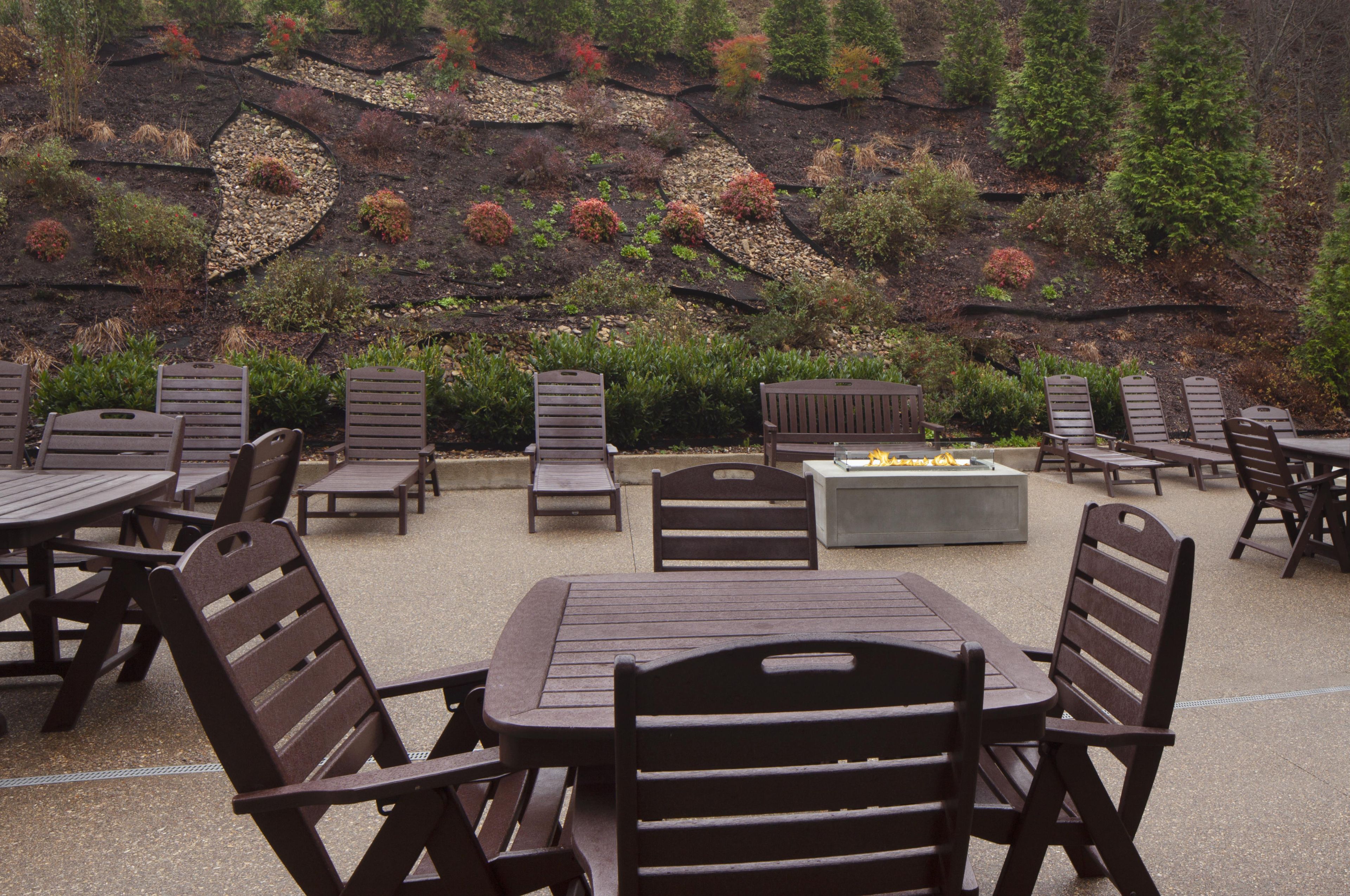An outdoor seating area with multiple tables and chairs, a fire feature, and a landscaped backdrop.