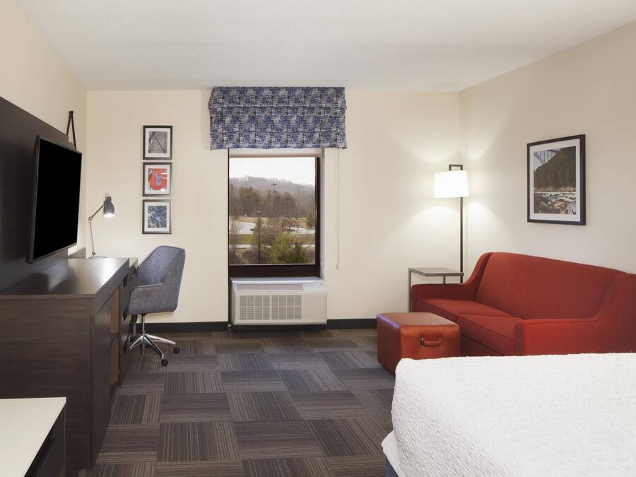 A hotel room with a bed, a desk with a TV, a chair, a red sofa, a small table, a lamp, and a window with a view of trees and a building.