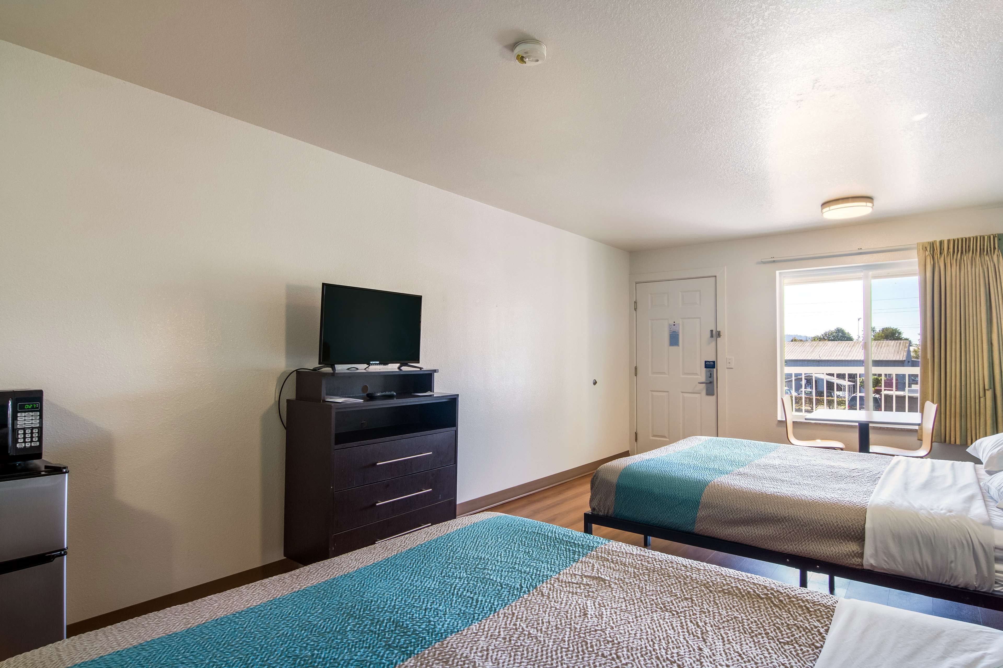 A hotel room with two beds, a TV on a dresser, a refrigerator, and a window with curtains.
