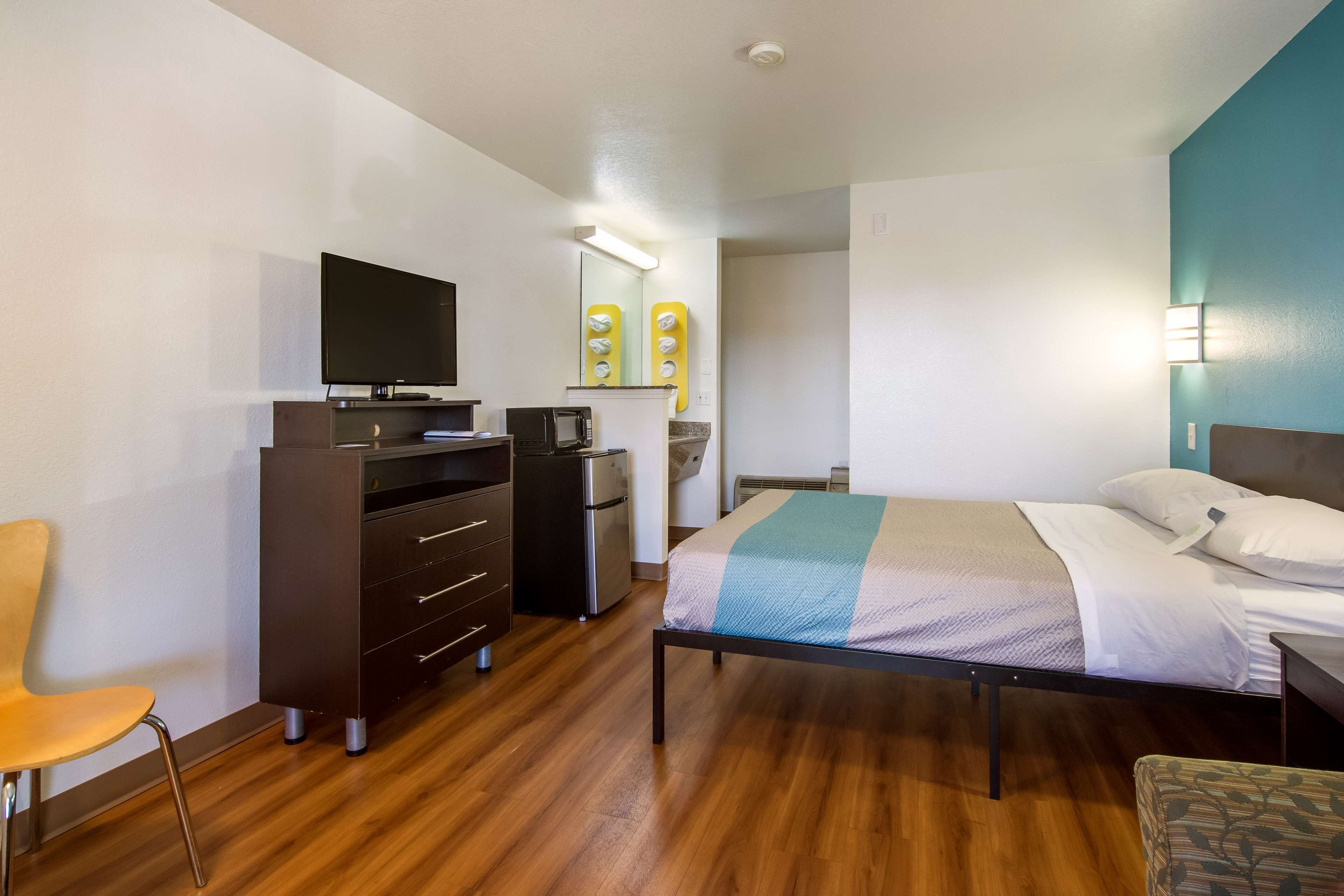 A hotel room with a bed, a desk with a computer, a chair, a small kitchenette, and a TV.