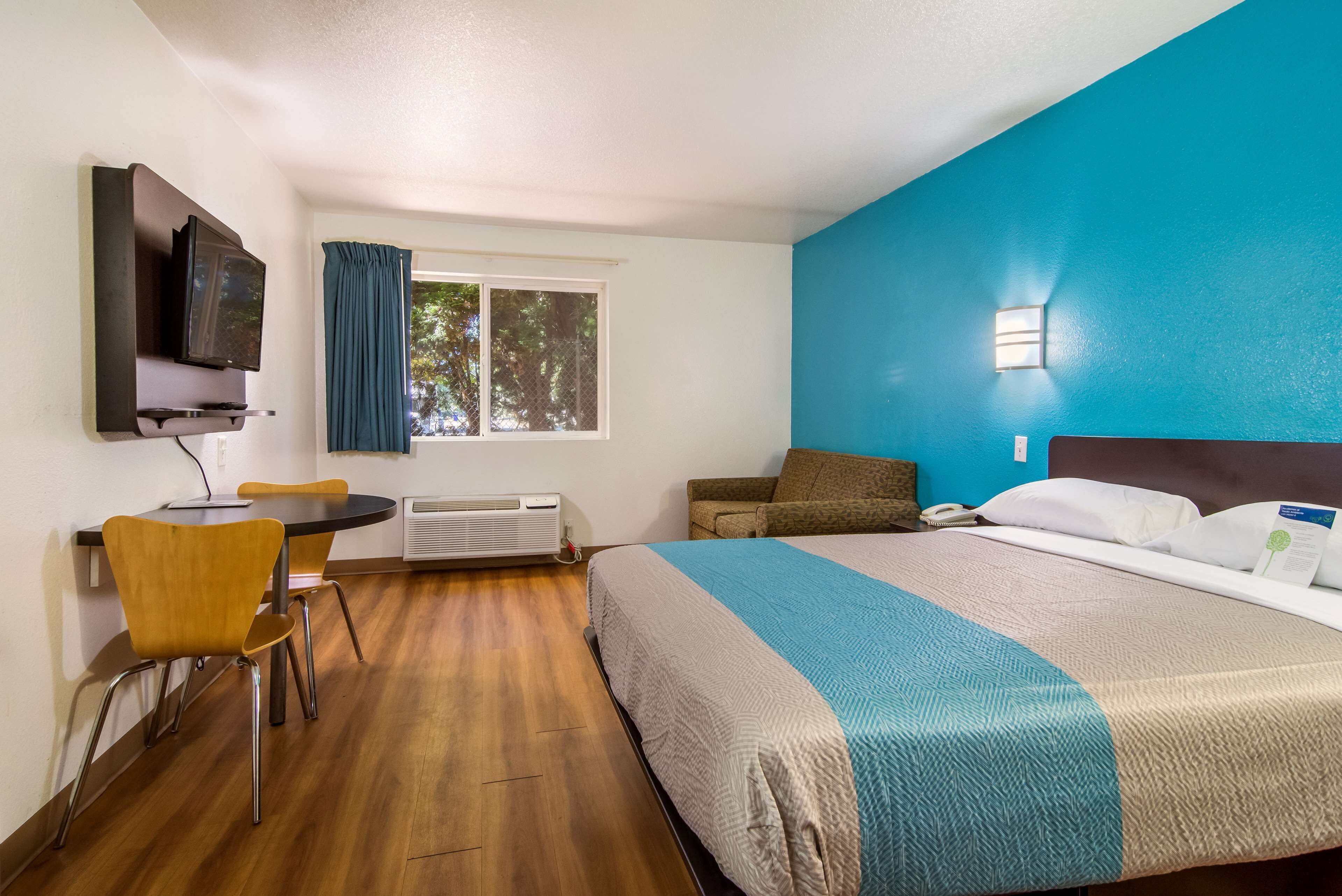 A hotel room with a bed, a desk with a chair, a television, a window with curtains, and a wall-mounted light.