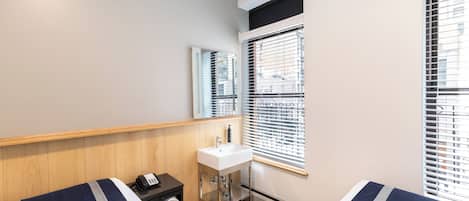 Une chambre d’hôtel avec deux lits, une table de chevet avec un téléphone, un petit lavabo et une fenêtre avec des stores.