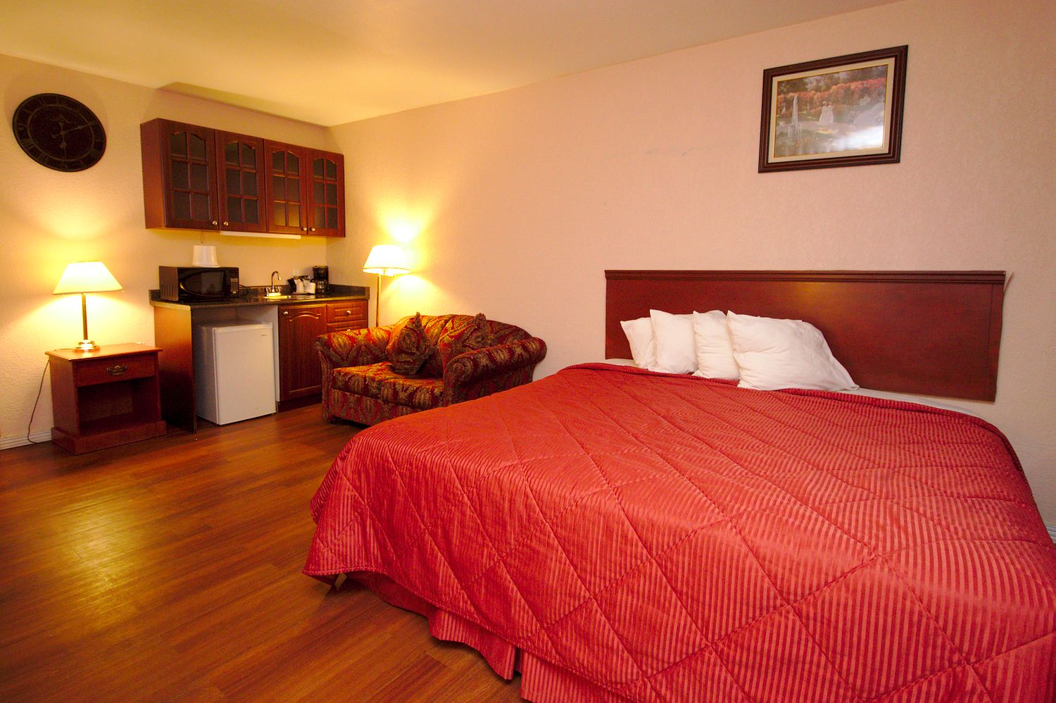 A hotel room with a bed, a chair, a small kitchenette, and a clock on the wall.