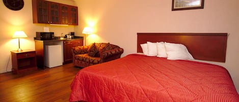 A hotel room with a bed, a chair, a small kitchenette, and a clock on the wall.