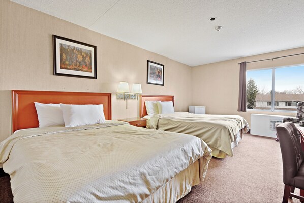 A hotel room with two beds, a desk, and a chair. There are two framed pictures on the wall, a window with curtains, and a ceiling light.